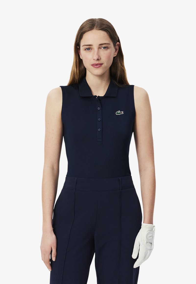 Polo sans manches bleu marine avec un col et une patte de boutonnage, arborant un petit logo de crocodile vert. Porte avec un pantalon bleu marine et un gant de golf blanc.