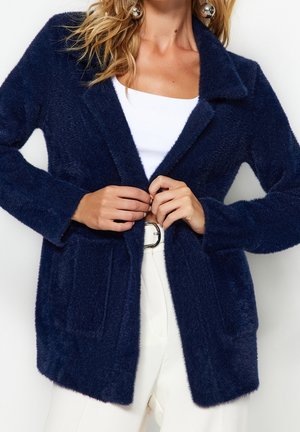 Vrouw draagt een marineblauwe, zacht uitziende cardigan over een wit shirt en witte broek, en maakt de voorkant met haar handen vast.