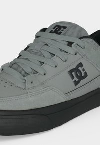 Gros plan d'une basket DC grise avec languette en mesh, lacets épais, superpositions en suède, semelle noire et logos sur le côté et la languette.
