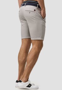 Shorts gris clair avec ourlets roulés, poches latérales et une poche arrière. Associés à des baskets blanches avec des accents noirs et une ceinture en nylon texturée bleu marine.
