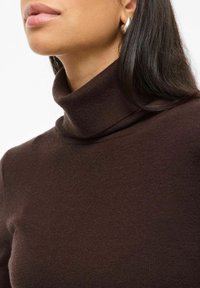 Maglione a collo alto marrone con un design aderente e colletto a coste. Texture liscia e maniche lunghe, abbinato a un orecchino a cerchio in argento.