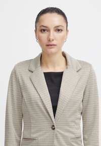 Beige checkered blazer with notch lapels, single button closure, and two side pockets, worn over a black top. Smooth, structured fabric.