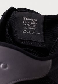Black sneaker interior with a fabric lining, featuring a stitched tag displaying a quote in white text. Smooth black accents visible.