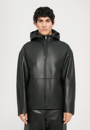 Young man wearing a black leather zip-up jacket with a hood, standing against a plain light gray background.