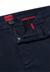 Navy blue trousers featuring a zip and button closure, red accents on the inside waistband, and a smooth texture. HUGO branded tag visible.