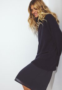 Jūrinės spalvos megztinis su aukštu kaklu, turintis sutrauktas rankoves, derinamas su juoda permatomą sijoną. Minkšta tekstūra ir laisvas kirpimas.