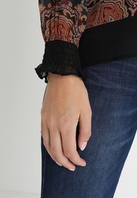 Polsino arricciato nero con maniche lunghe trasparenti a fantasia, abbinato a jeans in denim blu scuro. Focus sulla mano appoggiata sui jeans, evidenziando le texture.