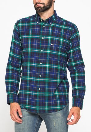 Uomo che indossa una camicia a quadri blu, verde e nera a maniche lunghe con jeans, in piedi su sfondo bianco.