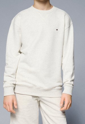 Enfant portant un sweat-shirt beige clair uni et un pantalon assorti sur un fond neutre, le sweat-shirt a un petit logo sur la poitrine.