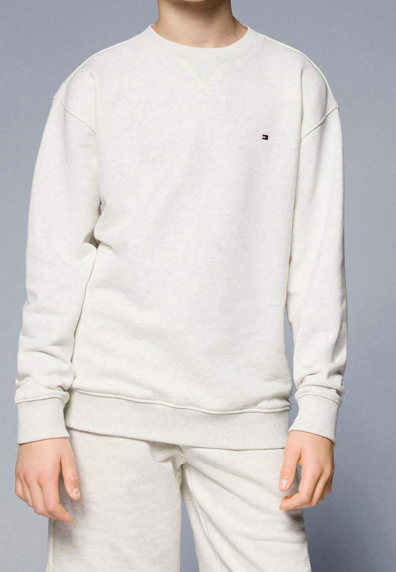 Enfant portant un sweat-shirt beige clair uni et un pantalon assorti sur un fond neutre, le sweat-shirt a un petit logo sur la poitrine.