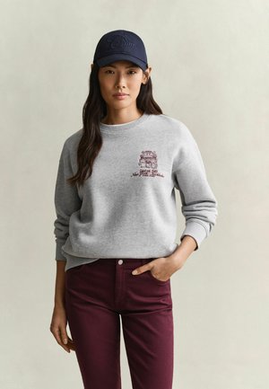 Jeune femme portant un sweat-shirt gris avec un emblème bordeaux, un pantalon bordeaux et une casquette de baseball bleu marine, debout avec une main dans la poche.
