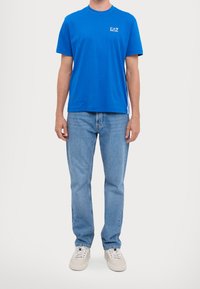 Modré bavlněné tričko s krátkým rukávem a kulatým výstřihem, s bílým logem "EA7" na hrudi. Kombinováno se světle modrými džíny a teniskami.