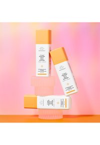 C-Luna Hydrabright Serum bottles with white bodies and orange caps, arranged over a pink background and a textured pink pedestal.