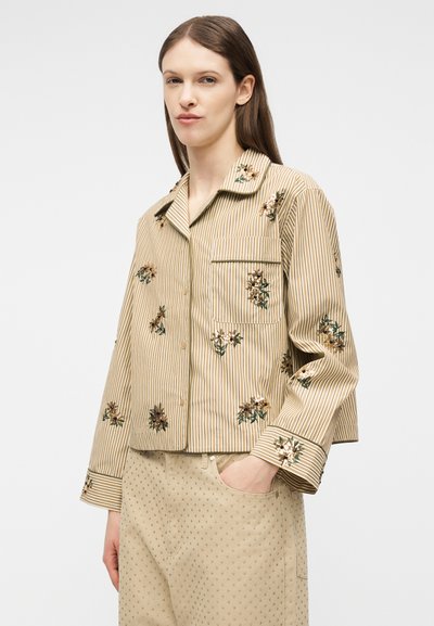 Γυναίκα με γαλάζιο ριγέ πουκάμισο με κεντήματα λουλουδιών και μπεζ παντελόνι με μικρό πουά μοτίβο, το χέρι στην τσέπη, ουδέτερο φόντο.