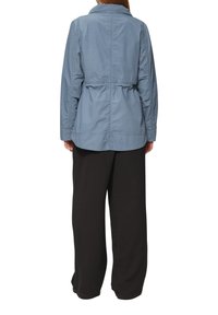 Giacca di tessuto azzurro chiaro con colletto, vita arricciata, maniche lunghe e taglio dritto; abbinata a pantaloni neri a gamba larga.