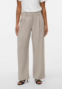 Ženska, oblečena v bež široke hlače z visokim pasom, belo majico, črne odprte pete in zlati prstani na prstih, stoji ob belem ozadju.