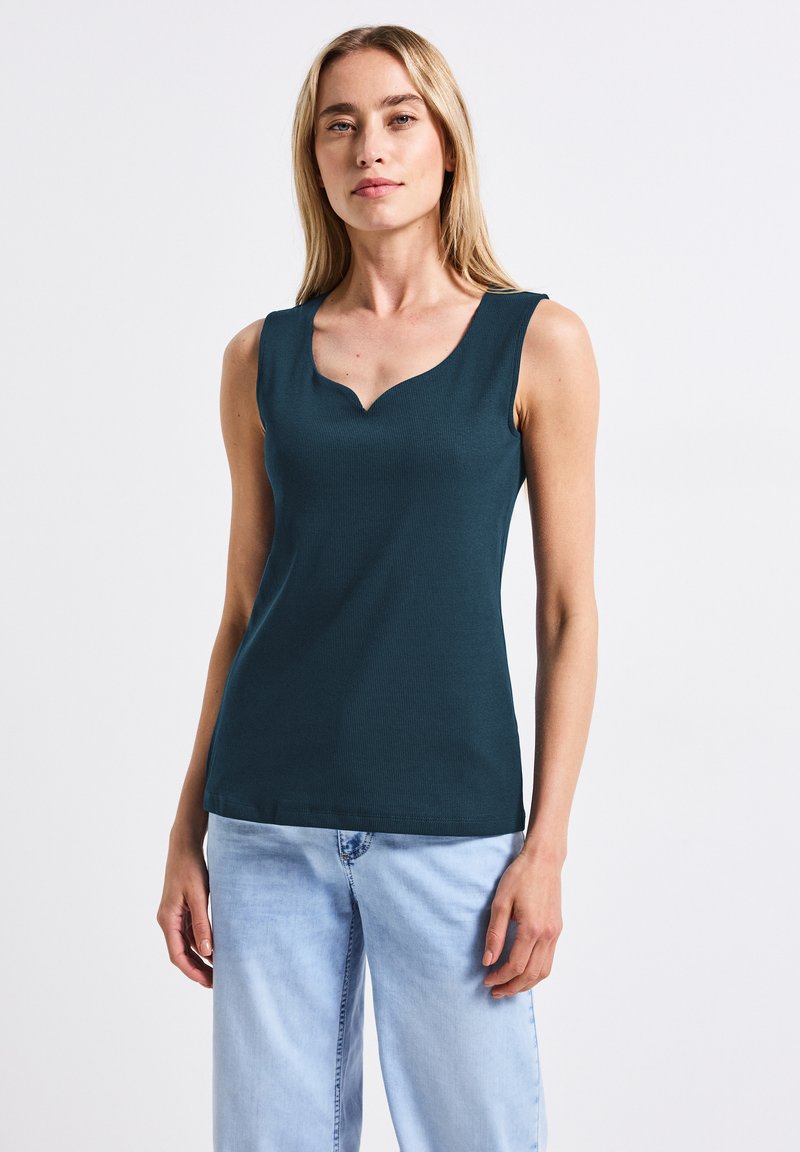 Top senza maniche in un verde scuro a coste, con scollatura ondulata, design aderente e texture liscia. Abbinato a jeans azzurri chiari.
