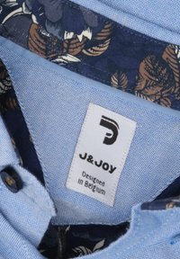 Blauw geblokt stof met een bloemenpatroon aan de binnenkant. Etiket toont "J&JOY" en "Ontworpen in België." Knopdetail zichtbaar.