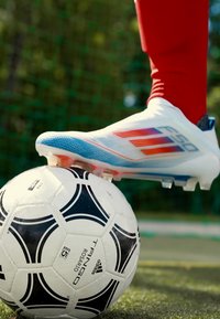 Botín de fútbol blanco con detalles en azul y rojo colocado sobre un balón de fútbol de patrón blanco y negro en un campo de hierba.