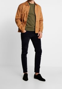 Chaqueta de algodón marrón sobre un sudadero verde, combinada con pantalones azul oscuro y zapatos negros. La chaqueta tiene un cierre de botones y bolsillos.