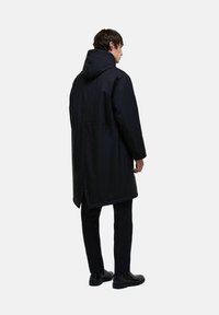 Hosszú fekete vízálló kabát kapucnival, egyenes szabású, oldalsó hasítékokkal és puha tapintású, fekete nadrággal és cipővel párosítva.