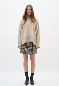 Beige stickad oversized tröja med hög krage, i kombination med en svart- och vitrutig kjol. Modellen bär svarta transparenta strumpor och spetsiga ballerinat.