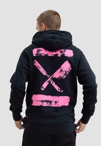 Mężczyzna w czarnej bluzie z kapturem, na plecach z nadrukiem dwóch skrzyżowanych różowych tasaków i tłuczka do mięsa, stojący tyłem na jednolitym tle.