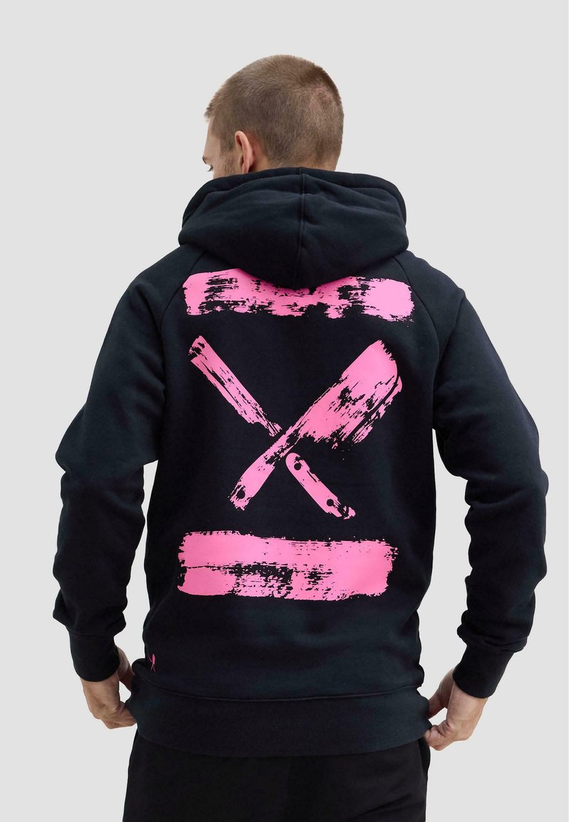 Mężczyzna w czarnej bluzie z kapturem, na plecach z nadrukiem dwóch skrzyżowanych różowych tasaków i tłuczka do mięsa, stojący tyłem na jednolitym tle.