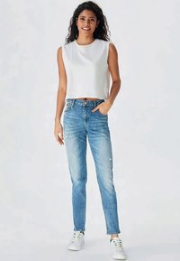 Balta saīsināta topa, zila augsta vidukļa džinsu bikses ar nodilušu dizainu, valkātas ar baltām kedām. Modelis stāv ar rokām kabatās.