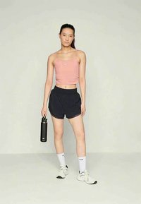 Młoda kobieta w różowym topie na ramiączkach, czarnych szortach, białych skarpetkach Nike i trampkach, trzymająca czarną butelkę na wodę, stojąca na jednolitym jasnym tle.