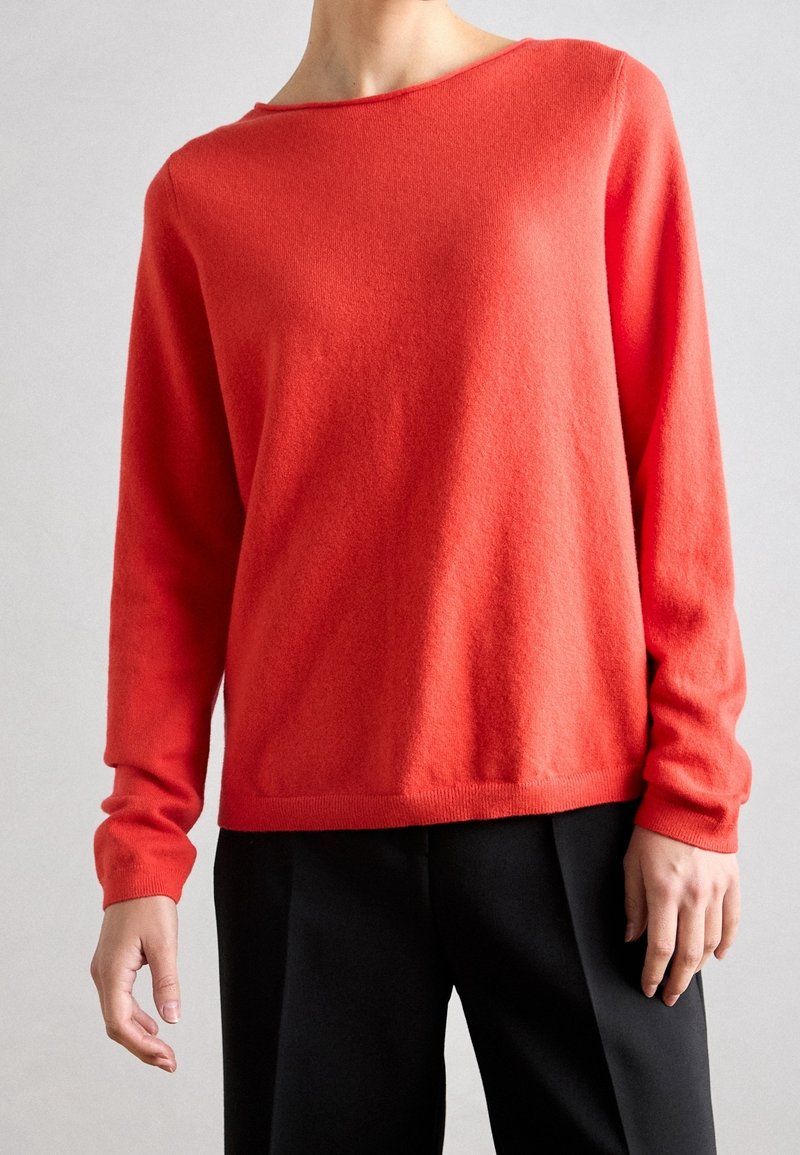 Personne portant un pull corail vif à manches longues avec une coupe décontractée et un pantalon noir sur un fond neutre.
