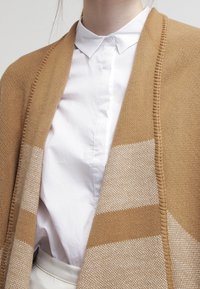 Bruine gebreide cardigan met een beige gestreept patroon, gedragen over een wit overhemd met kraag. Heeft een geribbelde rand en een zachte textuur.