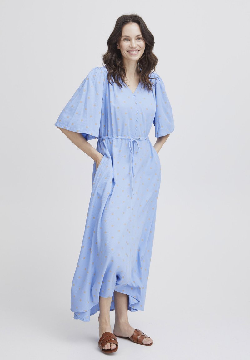 Himmelblaues knöchellanges Kleid mit kurzen Ärmeln, V-Ausschnitt, Kordelzug in der Taille, goldenen Pünktchen und seitlichen Taschen. Braune Sandalen runden den Look ab.