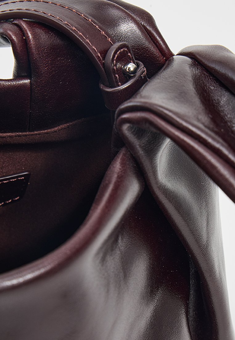Sac à main en cuir bordeaux foncé avec une texture lisse, une anse roulée et des détails de couture le long des bords, montrant une ouverture sur le dessus.