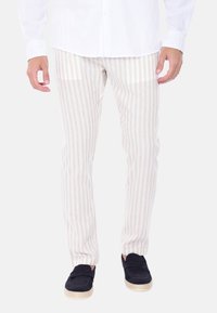 Pantaloni bianchi in lino con strisce verticali beige, dal taglio slim, caratterizzati da una texture leggera. Abbinati a scarpe scivolate scure.