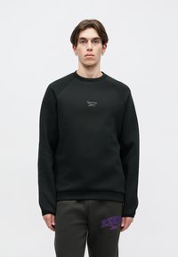 Persona joven vistiendo sudadera negra de Reebok y pantalones oscuros con el logo púrpura de Reebok, de pie contra un fondo claro y liso.