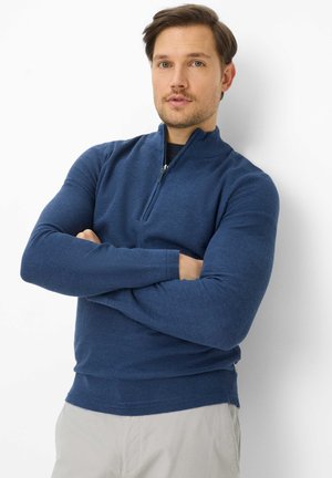 Man met gekruiste armen, gekleed in een blauwe trui met een kwartritssluiting en lichtgekleurde broek, staand tegen een witte achtergrond.