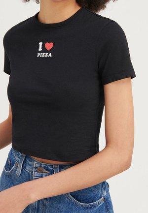 Vrouw draagt een zwart cropped T-shirt met de tekst "I [heart] PIZZA" en een blauwe spijkerbroek tegen een effen lichte achtergrond.