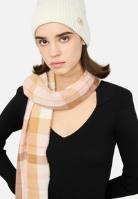 Cappello a cuffia bianco in maglia a coste con accento dorato. Sciarpa a quadri rosa e beige con bordi sfrangiati. Top nero con scollo a V e maniche lunghe.