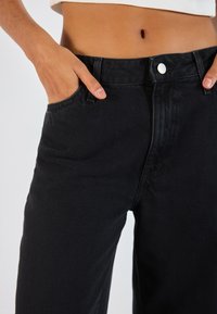 Personne portant un jean en denim noir avec une main dans la poche avant et un crop top blanc montrant le ventre.