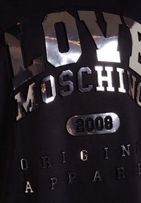 Černé tričko s metalicky stříbrným grafickým textem "LOVE MOSCHINO 2008" a "ORIGINAL APPAREL" v různých stylech a velikostech písma.
