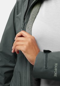 Donkergroene waterdichte jas met ritssluiting, fleece voering en verstelbare manchet met klittenband. De tekst "TEXAPORE" is zichtbaar op de mouw.
