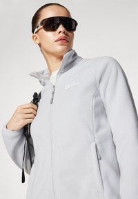 Giacca in pile grigia chiara con colletto alto, chiusura con zip e tasche laterali. Il modello indossa occhiali da sole neri e tiene una tracolla.