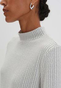 Haut texturé blanc avec un col haut, présentant un motif à carreaux. Porté avec une boucle d'oreille en argent brillant. Gros plan sur le profil latéral.