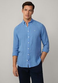Camicia di lino azzurro chiaro con colletto a polo, maniche lunghe arrotolate e un piccolo logo ricamato sul petto. Bottoni bianchi.