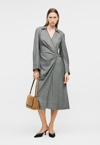 Γυναίκα με γκρι φόρεμα τύπου wrap με μακριά μανίκια, κρατώντας τσάντα από σουέντ σε ταμπα απόχρωση, και φορώντας μαύρες γόβες με μυτερή μύτη μπροστά σε λευκό φόντο.