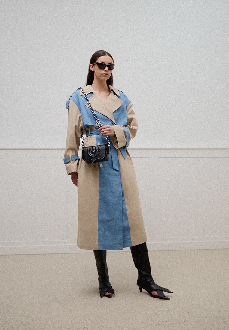 Manteau en beige et bleu denim avec un large col, sections en tissu dual, taille ceinturée, associé à un petit sac à main noir et des bottes noires élégantes.