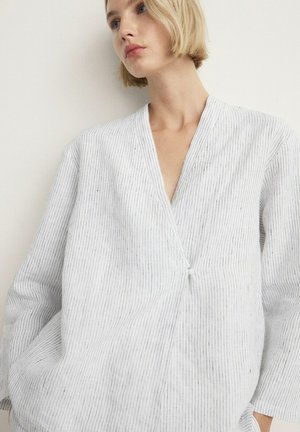 Jeune femme aux cheveux blonds courts portant un chemisier blanc à fines rayures, manches longues, col en V et fermeture nouée à l'avant.