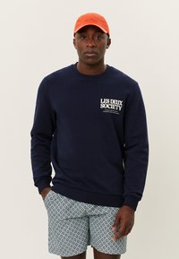 Sudadera marina con el logo "LES DEUX SOCIETY" en el pecho, combinada con pantalones cortos estampados que presentan diseños circulares, y una gorra naranja.