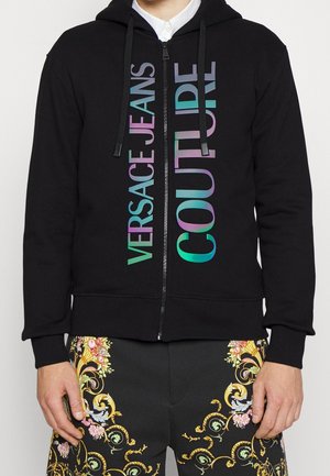 Sweat à capuche noir zippé avec un logo multicolore "Versace Jeans Couture" sur le devant, porté avec un pantalon noir présentant des motifs baroques dorés et floraux ornés.